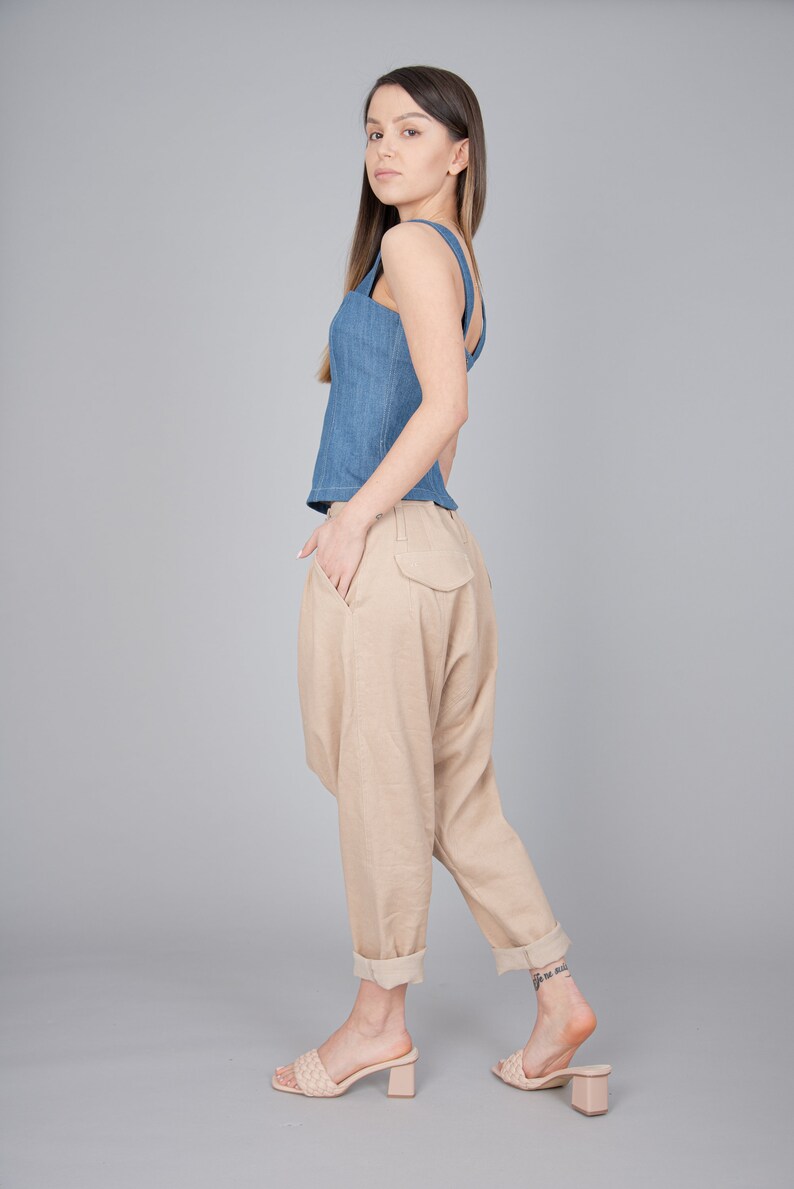 May include: A woman wearing a blue denim tank top and beige linen trousers. She is wearing beige heeled sandals and has her hair down.