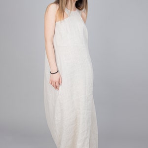 May include: A woman wearing a long, flowing, light beige linen dress. The dress has a halter neckline and a relaxed fit.