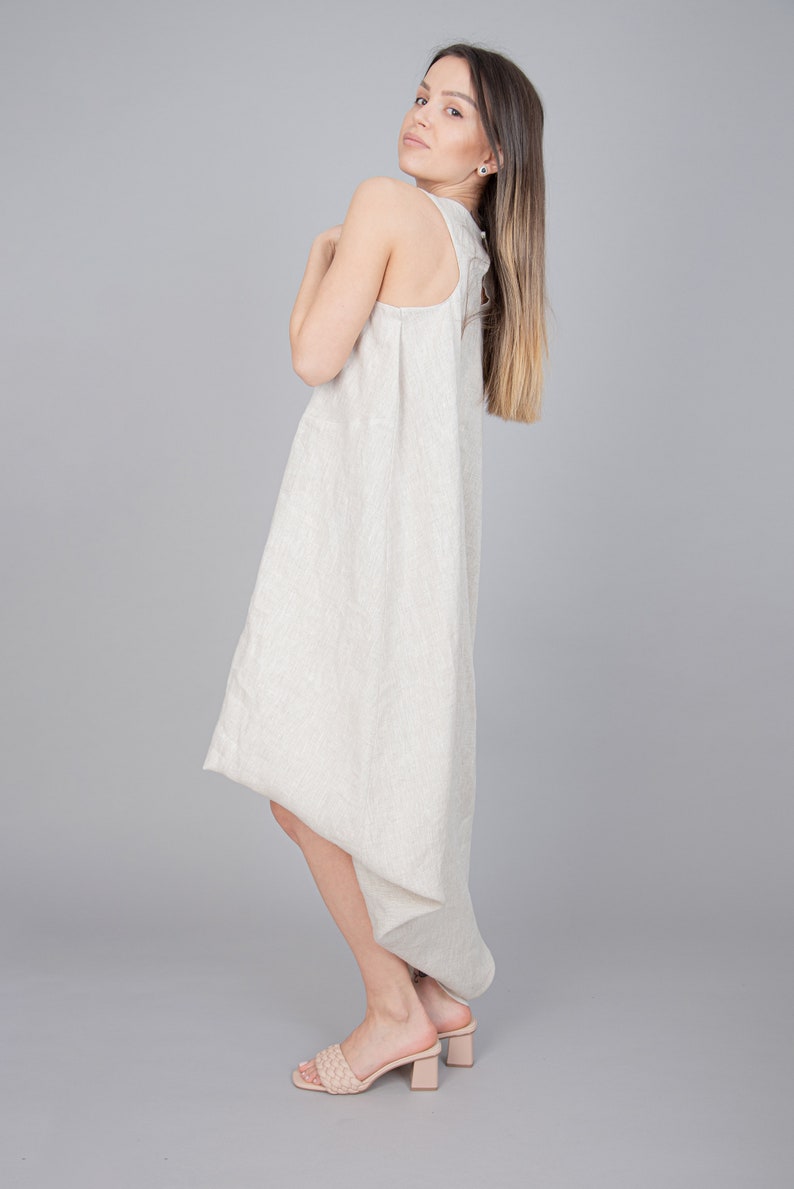 May include: A woman wearing a light beige linen dress with a high neckline and a flowing skirt. She is wearing nude heeled sandals.