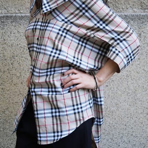 May include: A beige and black checked shirt with a long, asymmetrical hemline. The shirt has a relaxed fit and a collar.