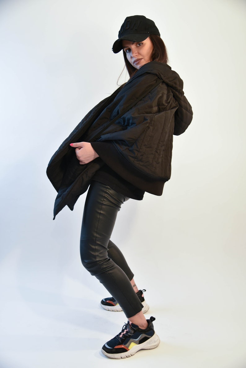 May include: A black quilted jacket with a hood, worn over black pants and sneakers. The person is wearing a black baseball cap with a white logo.