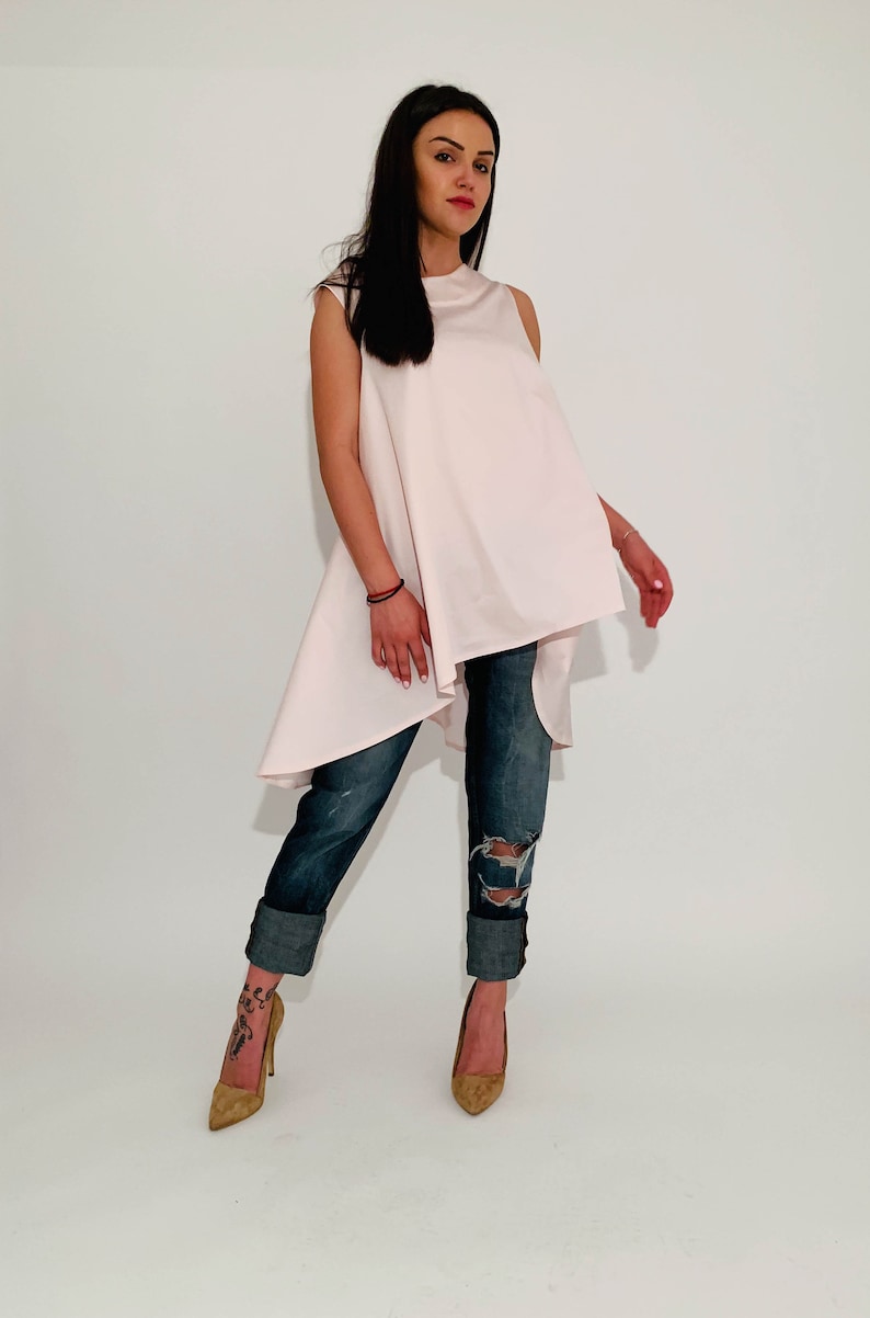 May include: A woman is wearing a light pink, sleeveless, asymmetrical top with a high-low hemline. She is also wearing blue jeans with distressed details and brown heeled shoes.