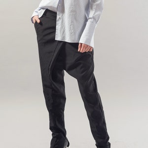 May include: A person wearing black trousers with a unique draped design and a light blue striped button-down shirt.