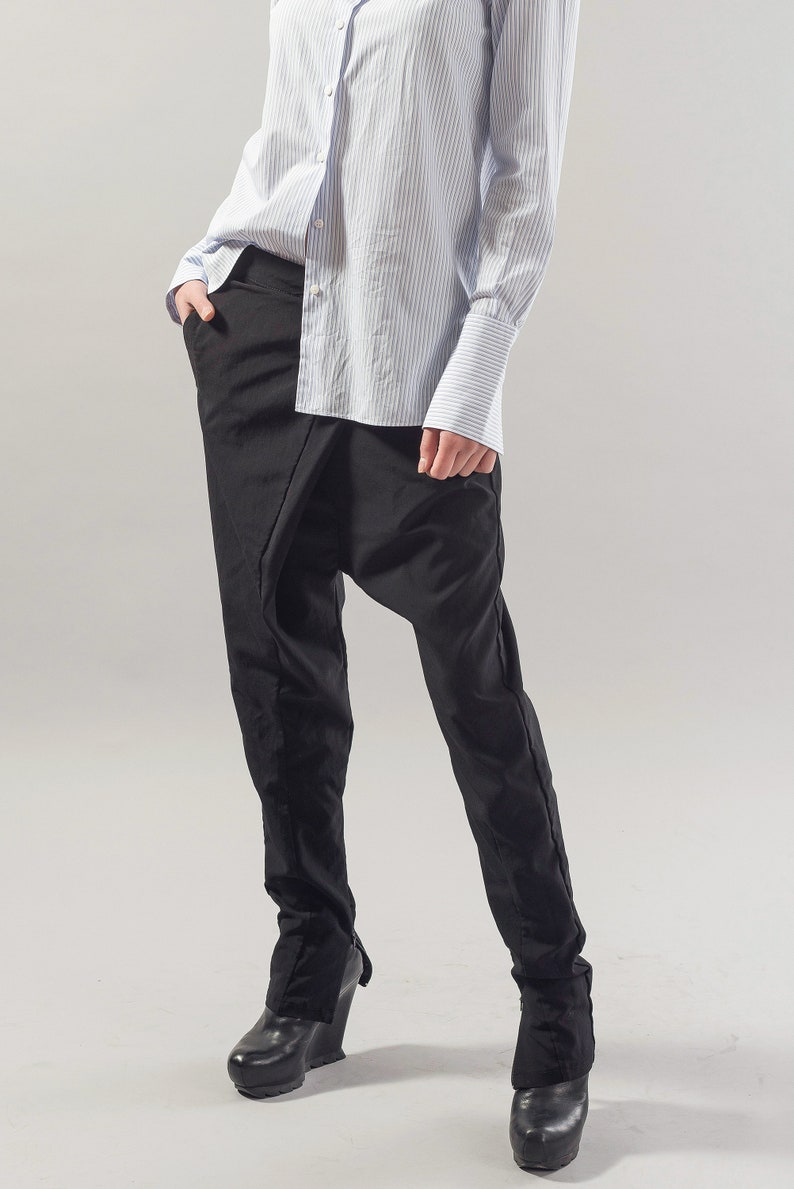 May include: A person wearing black trousers with a unique draped design and a light blue striped button-down shirt.