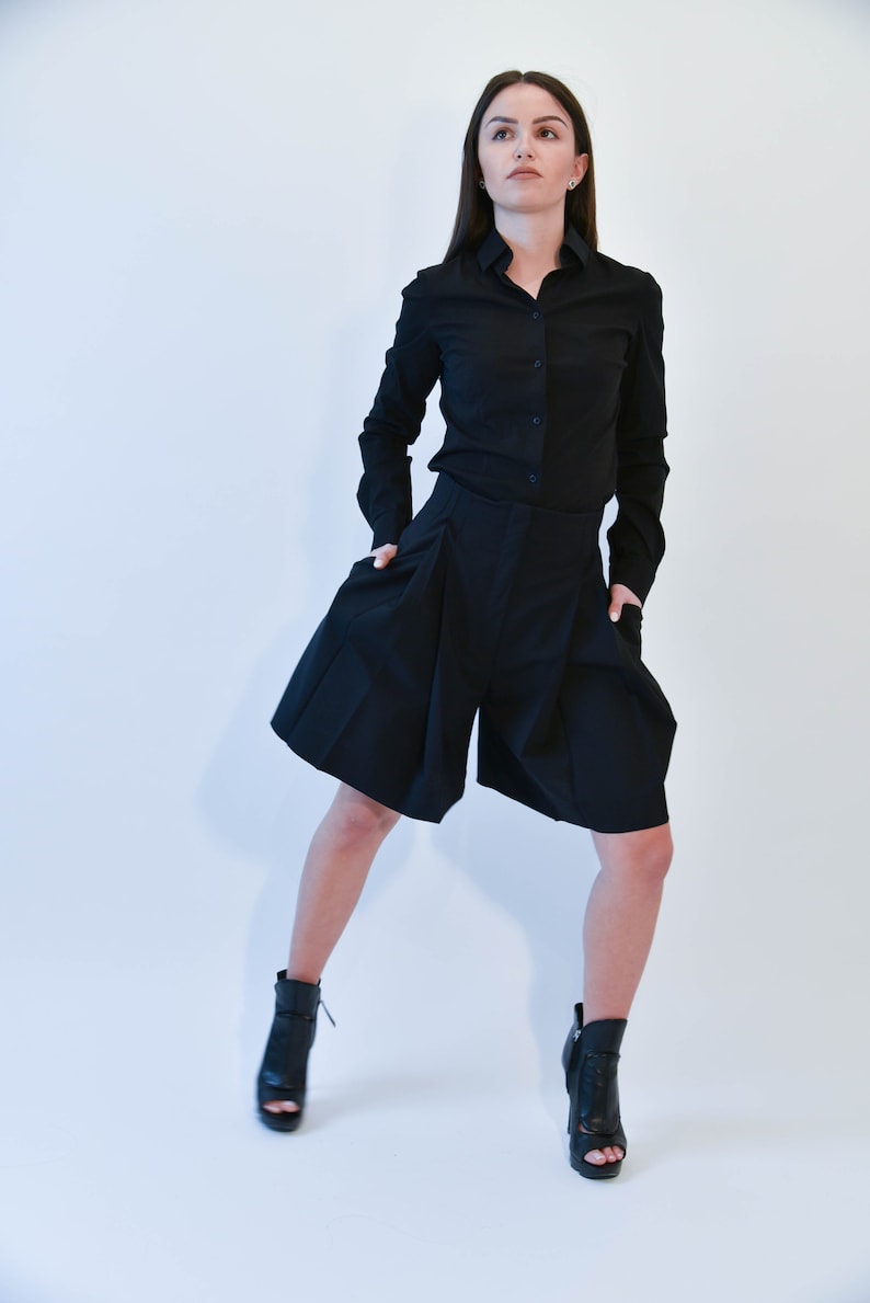May include: A woman wearing a black long-sleeved shirt and black shorts with pleats. She is wearing black heeled ankle boots.