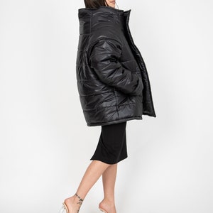 May include: A black puffer jacket with a high collar and a zipper closure. The jacket is worn by a person with long dark hair and is standing on a white background.