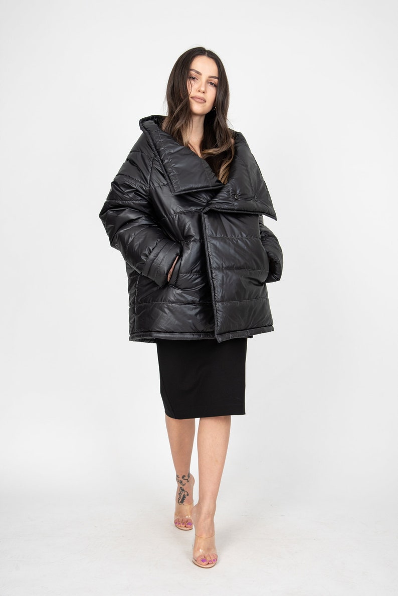 May include: A black puffer jacket with a large, asymmetrical collar. The jacket is quilted and has a shiny finish. The model is wearing a black skirt and clear heels.