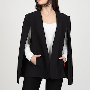 May include: A black cape blazer with a notched collar and two front pockets. The blazer is worn over a white top and black pants. The model is wearing the blazer with their hands in the pockets.