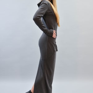 May include: A long, dark gray dress with a hood and long sleeves. The dress has a fitted silhouette and a slit up the side. The model is wearing black high-heeled boots. The background is a solid light gray.