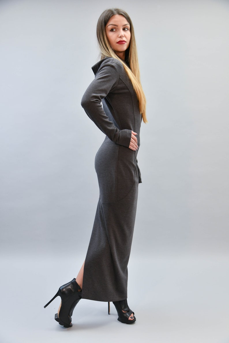 May include: A long, dark gray dress with a hood and long sleeves. The dress has a fitted silhouette and a slit up the side. The model is wearing black high-heeled boots. The background is a solid light gray.