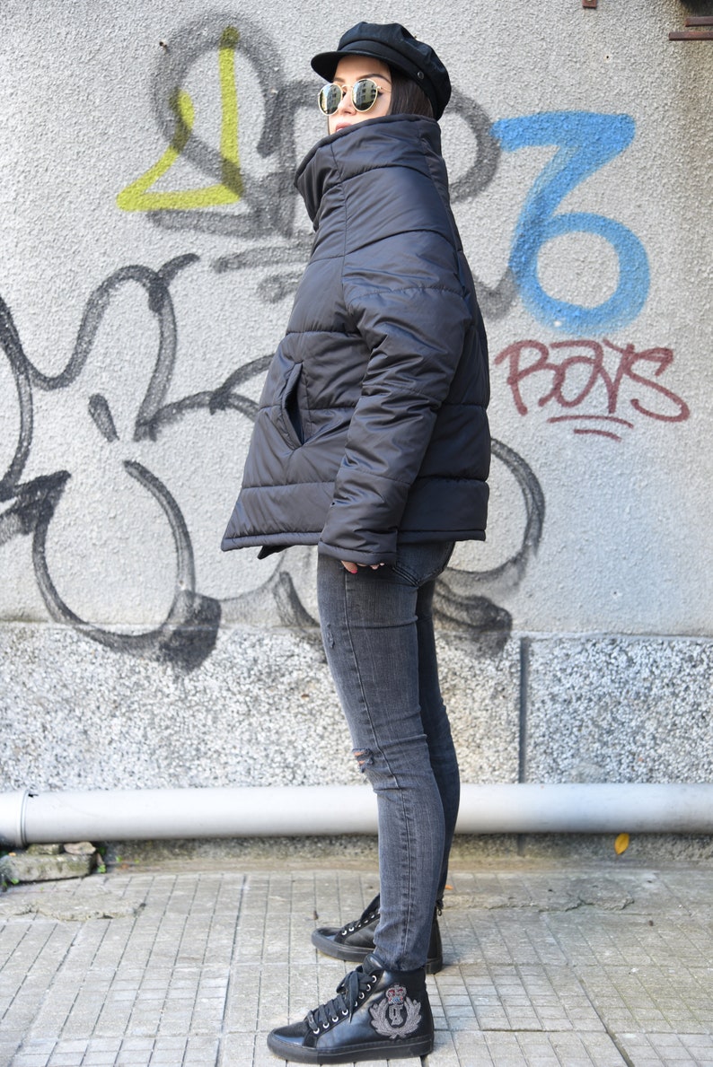 May include: A black puffer jacket with a high collar and a zipper closure. The jacket is worn by a person wearing gray jeans and black high-top sneakers with a crown emblem.