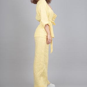 May include: A woman wearing a yellow linen pantsuit with a tie belt. She is standing with her right hand on her hip and her left hand holding the tie belt. She is wearing white trainers.