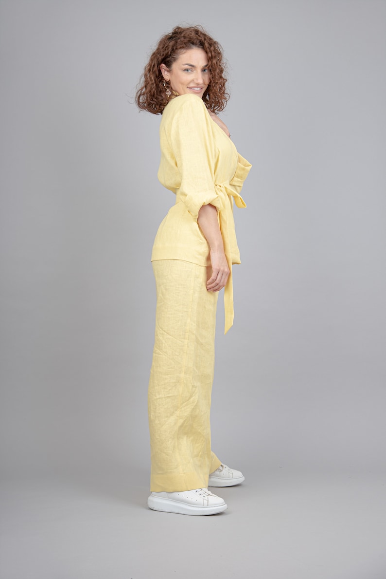 May include: A woman wearing a yellow linen pantsuit with a tie belt. She is standing with her right hand on her hip and her left hand holding the tie belt. She is wearing white trainers.