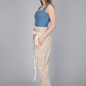 May include: A denim blue top with a square neckline and wide straps is paired with high-waisted, tan cargo pants. The pants feature large pockets and a matching fabric belt with a white rope tie. The outfit is completed with woven, light pink heeled sandals.