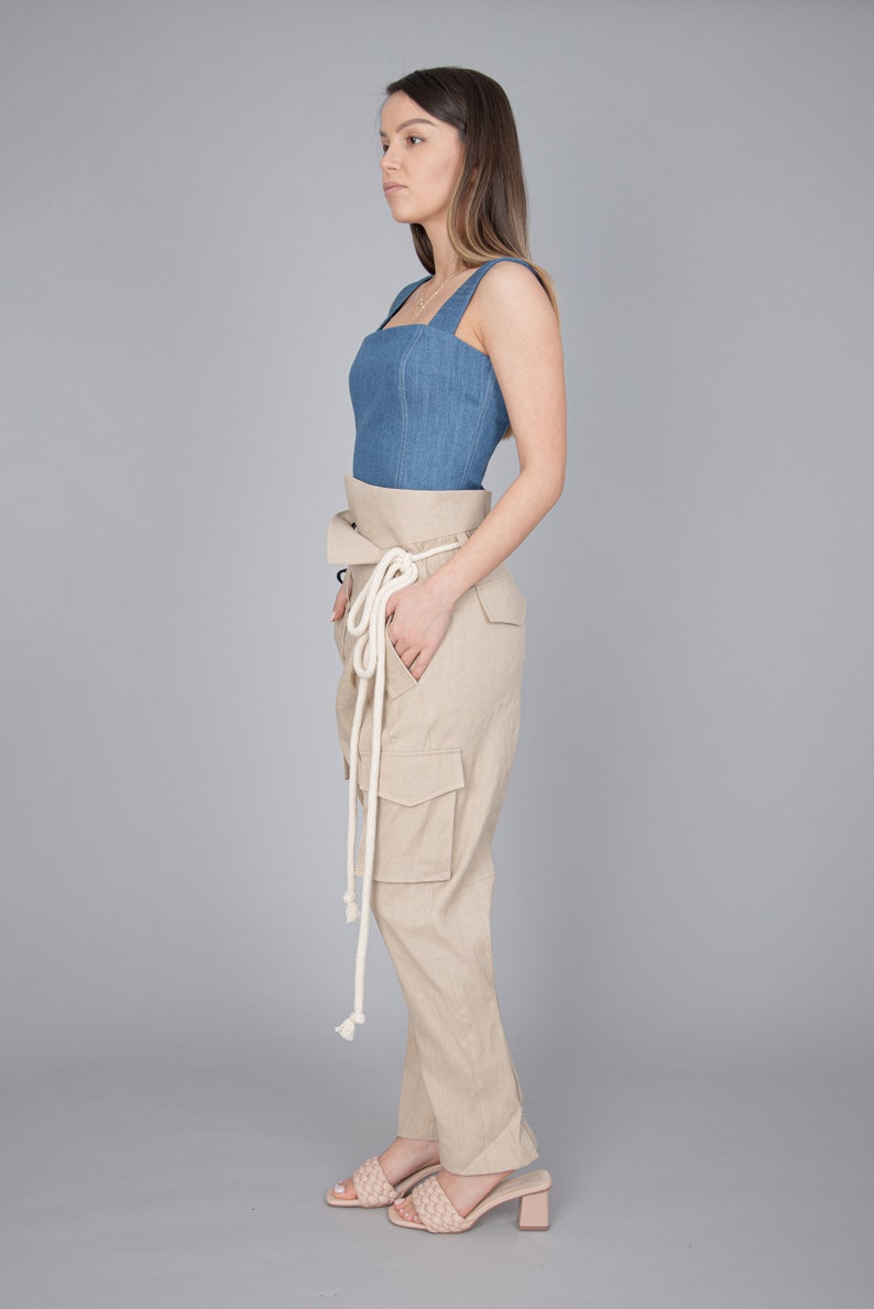 May include: A denim blue top with a square neckline and wide straps is paired with high-waisted, tan cargo pants. The pants feature large pockets and a matching fabric belt with a white rope tie. The outfit is completed with woven, light pink heeled sandals.