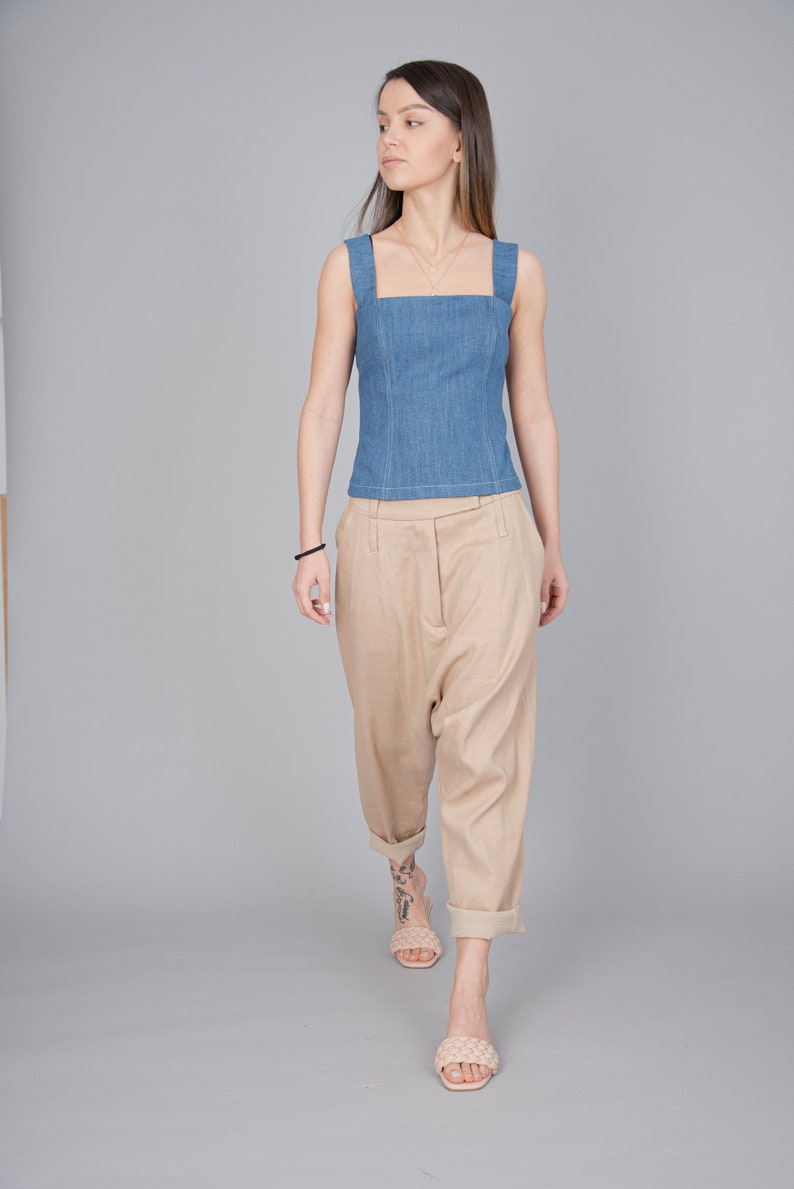 May include: A woman wearing a light blue denim sleeveless top and beige wide-leg trousers. She is wearing beige sandals and has a gold necklace.