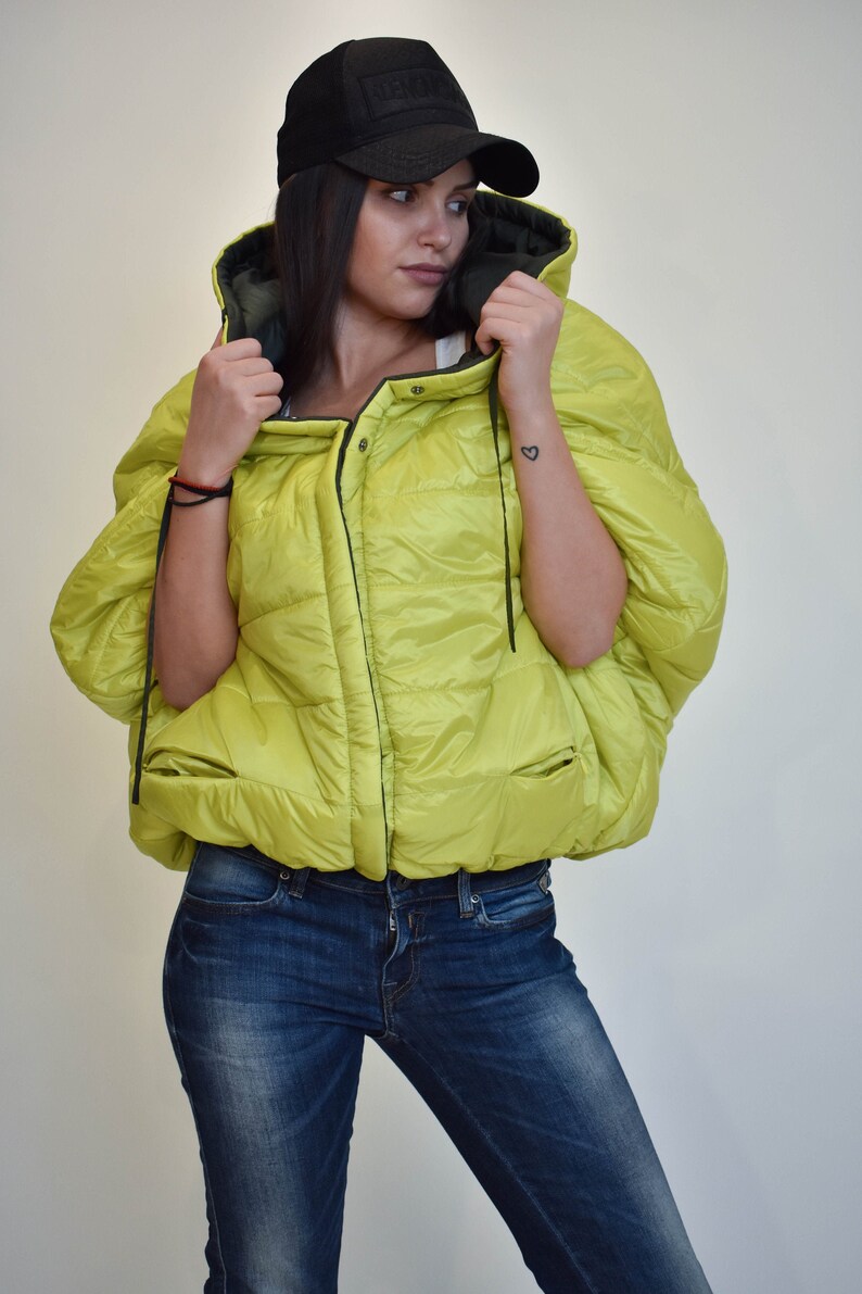May include: A lime green puffer jacket with a black zipper and a black hood. The jacket has a relaxed fit and a short length. The model is wearing blue jeans and a black baseball cap.
