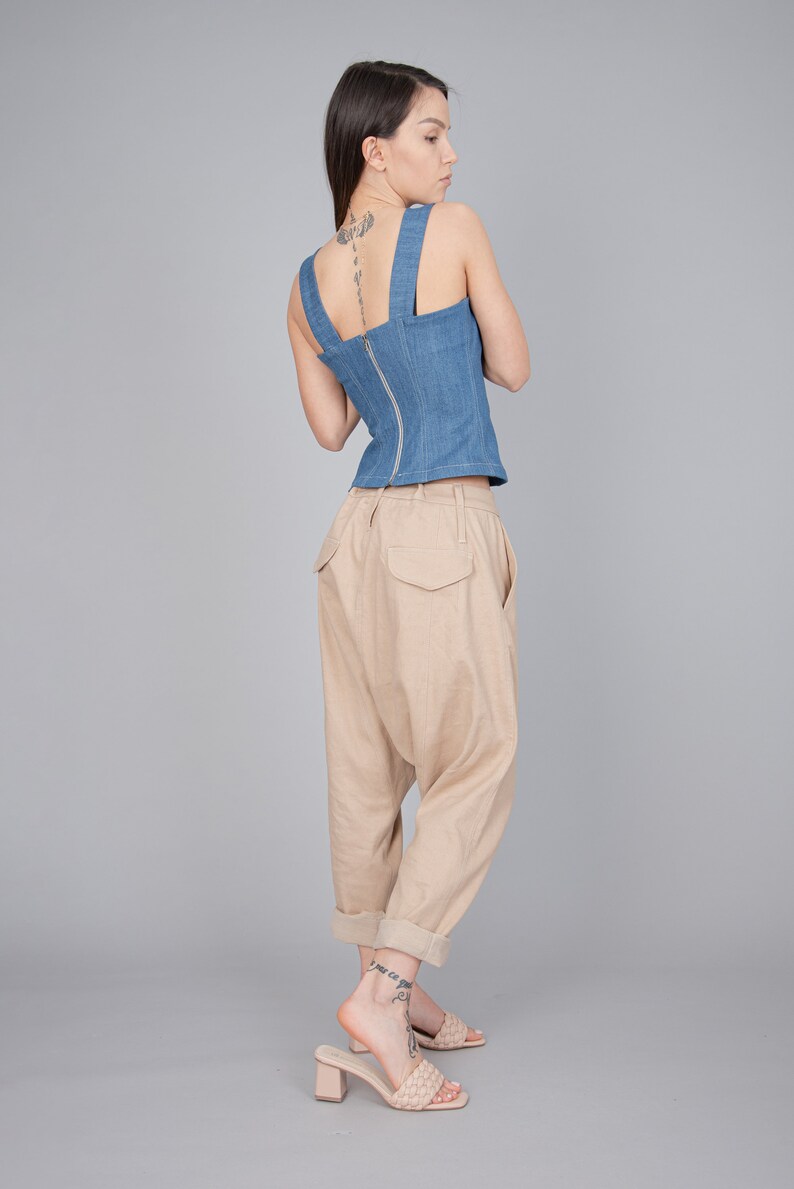 May include: A woman wearing a blue denim corset top with a zip down the back and beige, loose-fitting trousers. She is wearing beige heeled sandals with a woven strap.