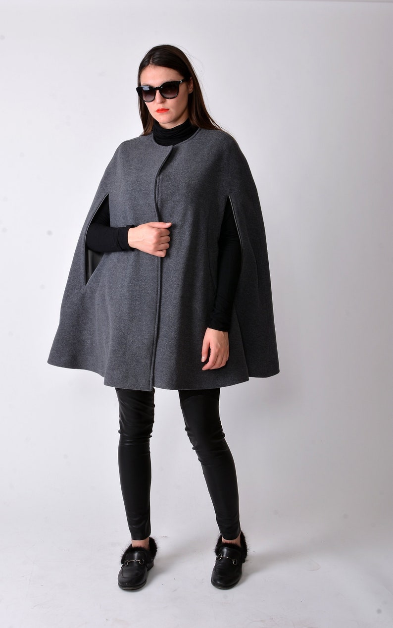 May include: A gray wool cape with a zipper closure and black leather trim. The cape is worn over a black turtleneck and black leggings. The person is wearing black loafers with fur trim.