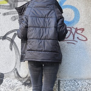 May include: A black puffer jacket with a high collar and a zipper closure. The jacket is worn over a pair of dark wash jeans.
