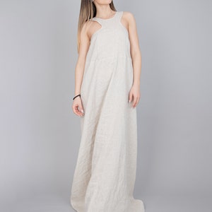 May include: A woman wearing a long, beige linen dress with a sleeveless design. The dress has a relaxed fit and a flowing silhouette.