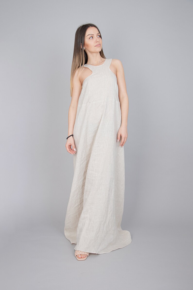 May include: A woman wearing a long, beige linen dress with a sleeveless design. The dress has a relaxed fit and a flowing silhouette.