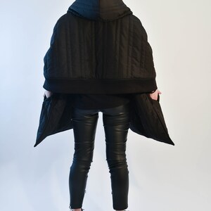 May include: A black quilted hooded jacket with a zipper closure and a black baseball cap with the word "SQUARED" on the front. The jacket is worn over black pants and black sneakers.