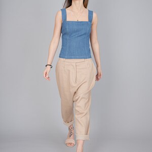 May include: A woman wearing a light blue denim tank top and beige linen trousers. She is wearing a gold necklace and has a tattoo on her ankle. She is walking towards the camera.
