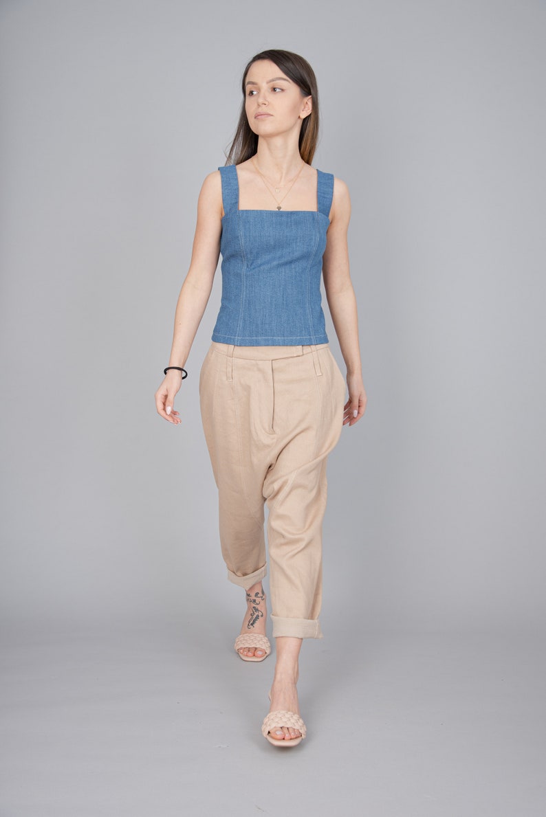 May include: A woman wearing a light blue denim tank top and beige linen trousers. She is wearing a gold necklace and has a tattoo on her ankle. She is walking towards the camera.