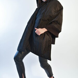 May include: A black quilted jacket with a hood and a black baseball cap. The jacket has a long, flowing design and is worn over black pants.