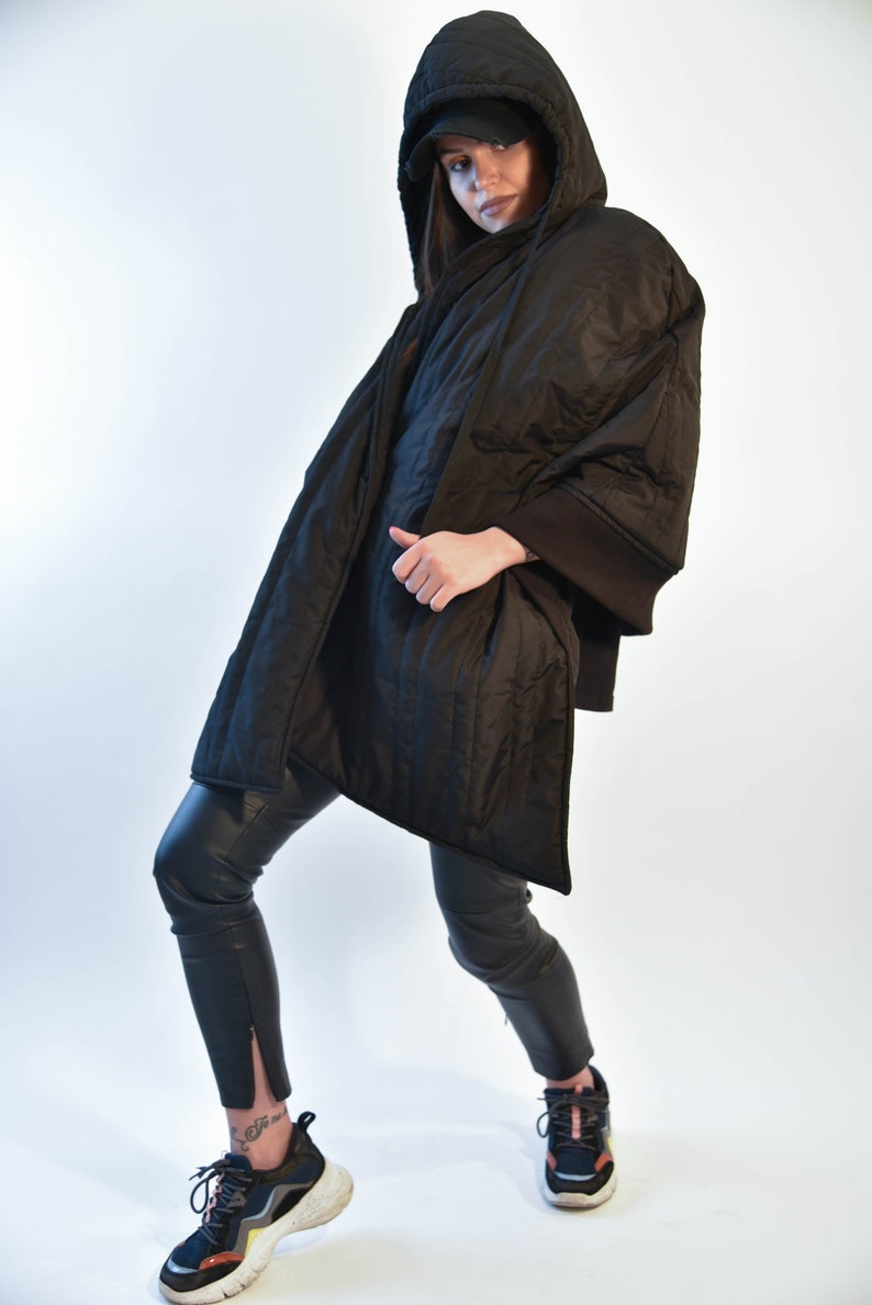 May include: A black quilted jacket with a hood and a black baseball cap. The jacket has a long, flowing design and is worn over black pants.