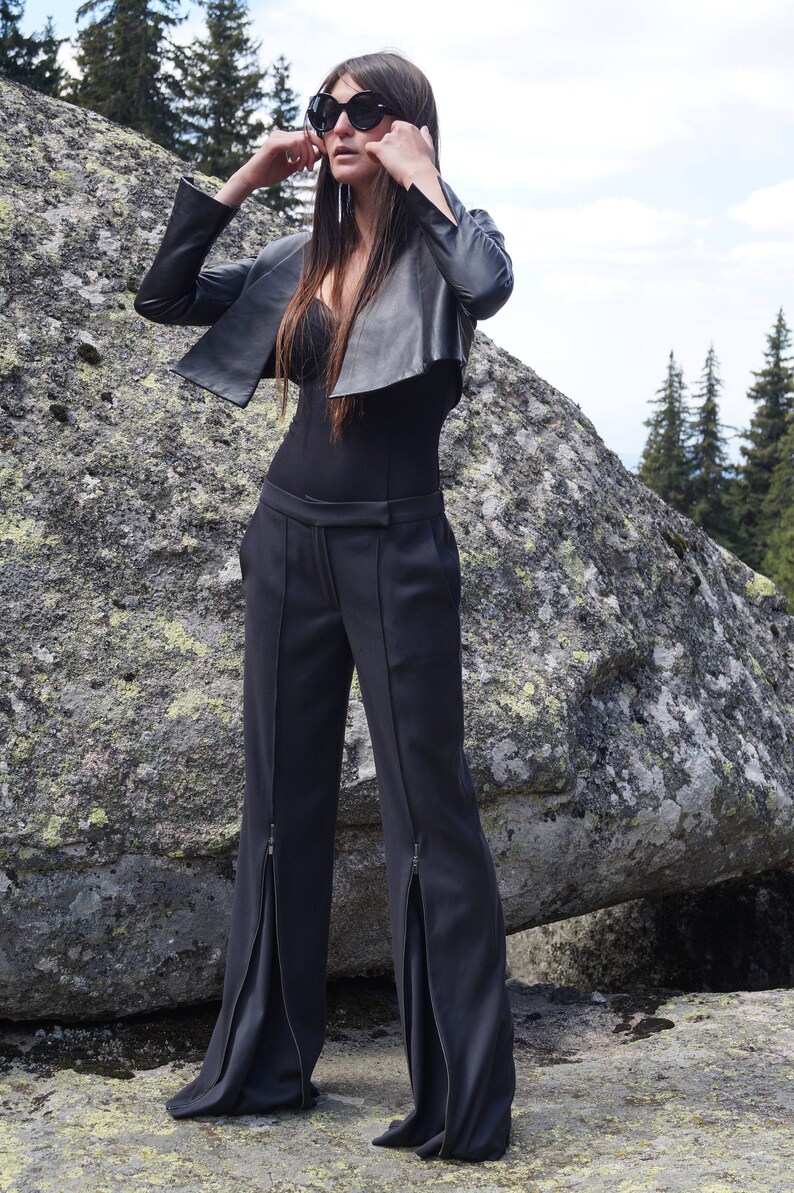 May include: A woman wearing a black leather jacket, black pants with zippers on the sides, and black sunglasses. She is standing on a large rock.