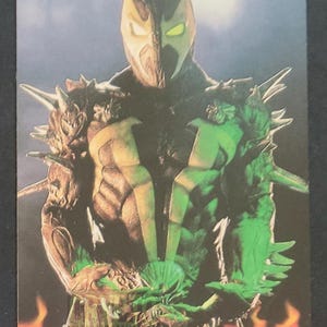 May include: A Spawn trading card featuring the character in a black and green costume with a mask. The card has a fiery border and the word "Spawn" is visible. The card also has the text "Collectibles Blockchains Local Comic Shop".
