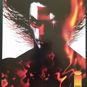 May include: Comic book cover art for HellShock #1, featuring a fiery red and black background with a character's face in the center. The character has a white face with a red cross painted on their forehead and glowing red eyes. The text "HellShock" is in large, bold letters at the top of the cover. The text "Jae Lee" is at the bottom of the cover.
