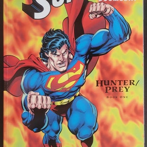 May include: A comic book cover featuring Superman in a red and blue suit flying through a fiery orange background. The title of the comic is "Superman Doomsday: Hunter/Prey Book One".