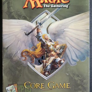 Puede incluir: Una caja de juego básico de Magic: The Gathering con un guerrero ángel de alas blancas que sostiene una espada y un escudo. La caja es verde y dorada con el texto "Magic: The Gathering" y "Core Game" en ella. El texto "Includes Premium Card and Magic Online" y "Two-Player Trading Card Game" también está en la caja.
