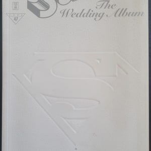 May include: A grey comic book cover for "Superman: The Wedding Album". The cover features the Superman logo and title in embossed lettering. The DC Comics logo is in the upper left corner, with issue details. A shop logo is at the bottom.