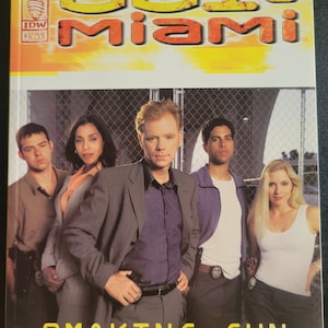 May include: A comic book cover featuring the title "CSI: Miami" and the words "Smoking Gun". The cover shows five characters, including a man in a suit, a woman in a white shirt, and a man in a blue shirt. The background is a metal fence.