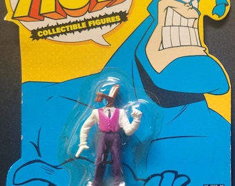 May include: Collectible action figure of The Tick, packaged on a yellow and blue card. The figure is of a man in a suit with a book for a head. The packaging reads "The Tick Collectible Figures."