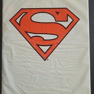 May include: A white plastic bag with a red and black Superman logo printed on the front. The bag has the text "THE MAN OF STEEL" printed on the bottom.
