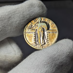 May include: A silver 1929 Standing Liberty quarter with a gold patina. The coin features the image of Liberty holding a torch and a branch of olive leaves. The inscription "IN GOD WE TRUST" is visible on the coin.