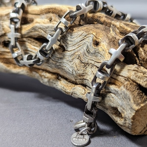 May include: A silver chain necklace with a cross design. The chain is thick and has a textured finish. The necklace is lying on a piece of driftwood.