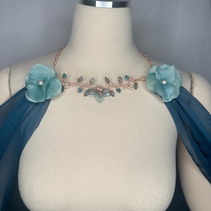 May include: A rose gold wire necklace featuring two large teal fabric flowers with central white pearls, accented by small teal beads, clear crystals, and leaf-shaped charms, connected to flowing teal fabric drapes on each side of the neck.