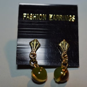 Könnte beinhalten: Ein Paar goldfarbene Ohrringe mit grünen und braunen Steinen. Die Ohrringe befinden sich auf einer schwarzen Karte mit dem Text "FASHION EARRINGS".