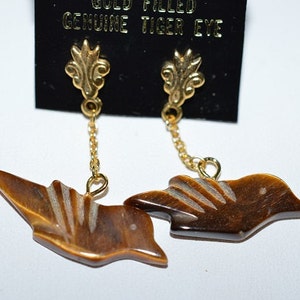 Pode incluir: Brincos de ouro com uma pedra de olho de tigre em forma de pássaro. Os brincos estão em um cartão preto com o texto "Gold Filled Genuine Tiger Eye".