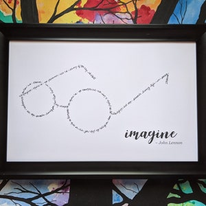 May include: A framed print with a black frame and a white background. The print features a pair of glasses made of the words "Imagine" by John Lennon. The words are written in black ink.