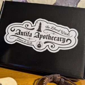 May include: A black box with a white label featuring the text "Antifa Apothecary: Potions & Spells" in an ornate font. The label also includes an illustration of a witch and a potion bottle. The box is on a wooden surface.