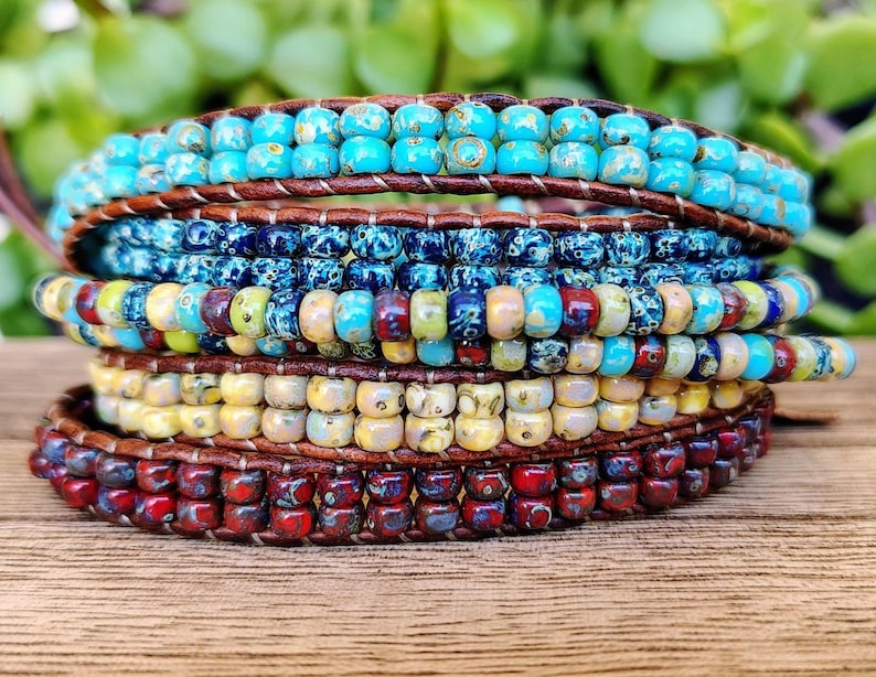 Op de afbeelding: Een wikkelarmband met meerdere strengen, bestaande uit rijen kleine, ronde, veelkleurige glaskralen die op een donkerbruin leren koord zijn genaaid. De kralen vertonen een kleurverloop van turkoois, diepblauw, geel, cr&egrave;me en donkerrood.