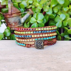 May include: A multi-strand wrap bracelet featuring rows of small, colourful glass beads in shades of red, lime green, and blue, woven onto brown leather cords. The bracelet is secured with a bronze-toned metal owl-shaped button closure.