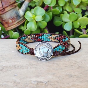 May include: A wrap bracelet featuring multi-coloured glass beads in shades of turquoise, red, and yellow, secured by a silver-toned United States five cents buffalo nickel coin button and brown leather cord ties.